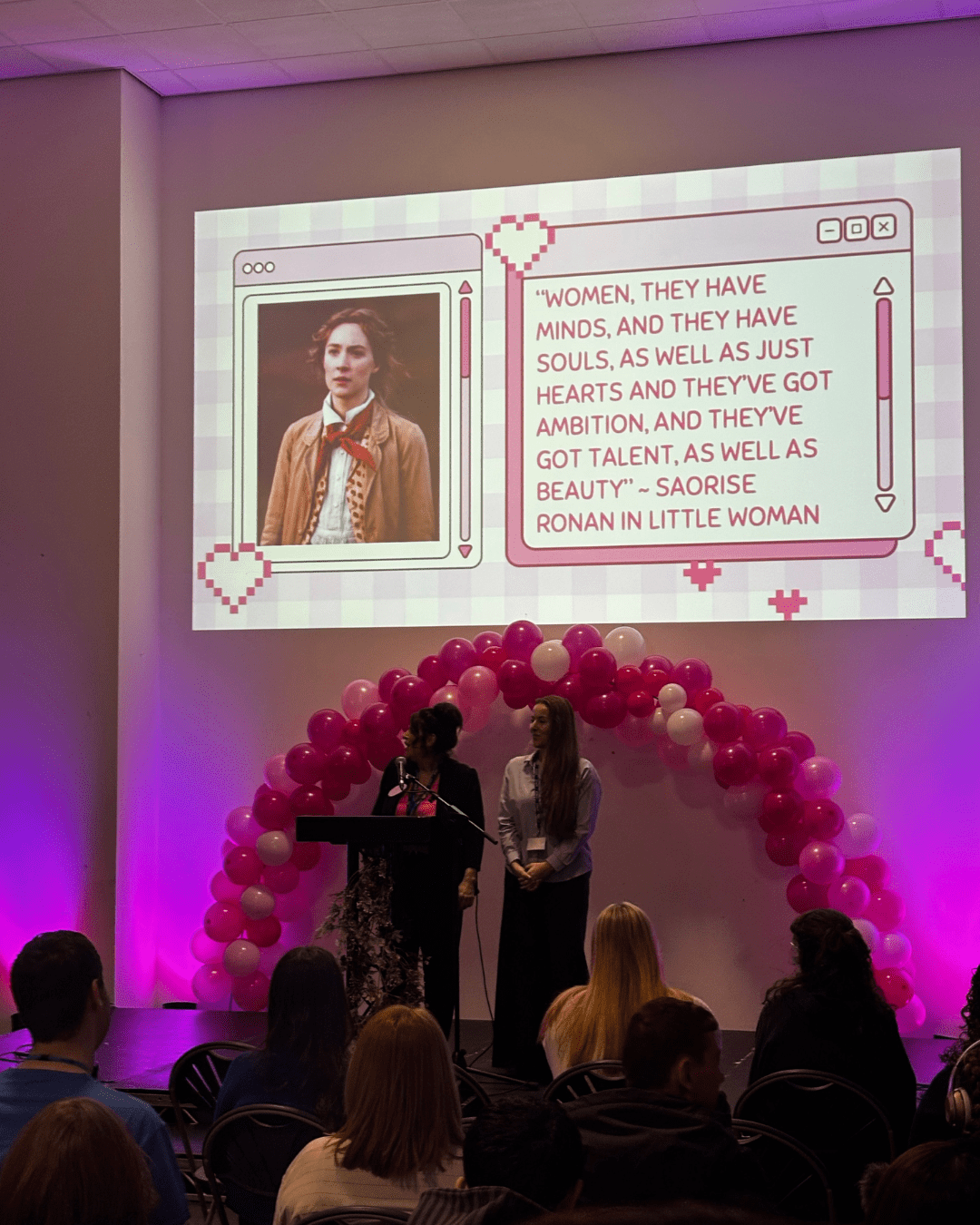 A speaker at the Celebrating Women event, there is a quote behind her from the film Little Women which reads: Women, they have minds, and they have souls, as well as just hearts and they've got ambition, and they've got talent, as well as beauty